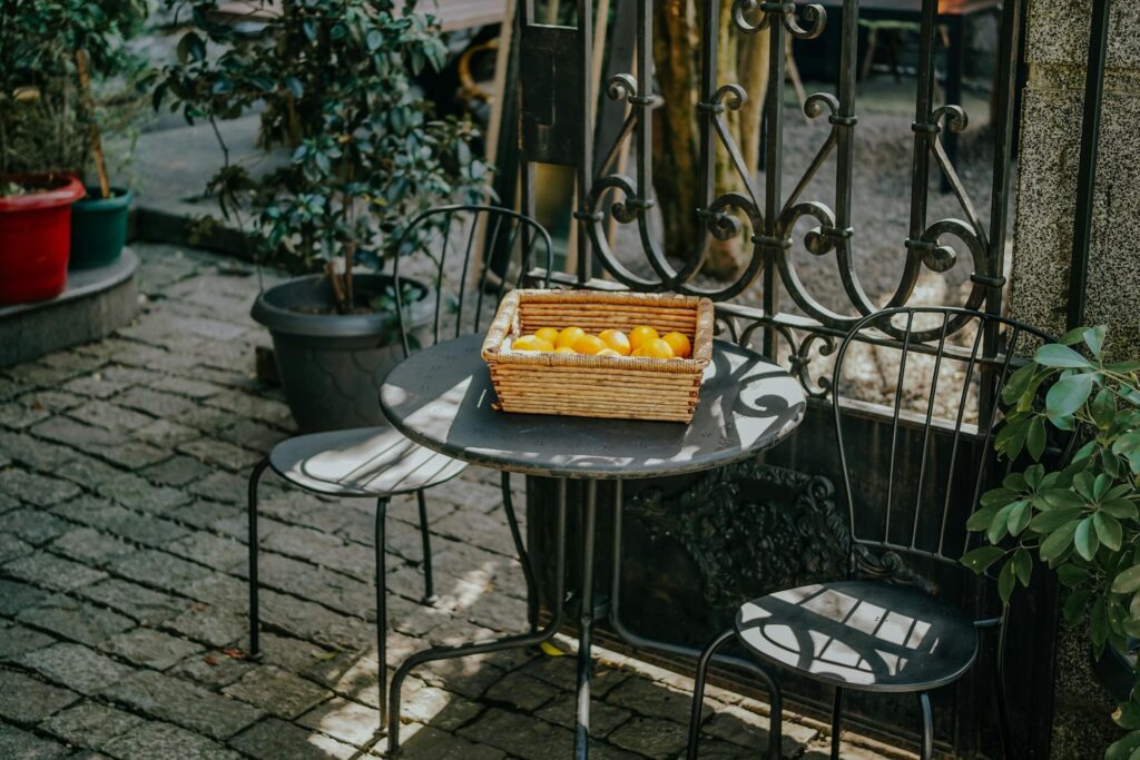 Urokliwy mały dziedziniec z metalowym stoliczkiem, krzesłami i koszem żółtych cytryn w otoczeniu zieleni