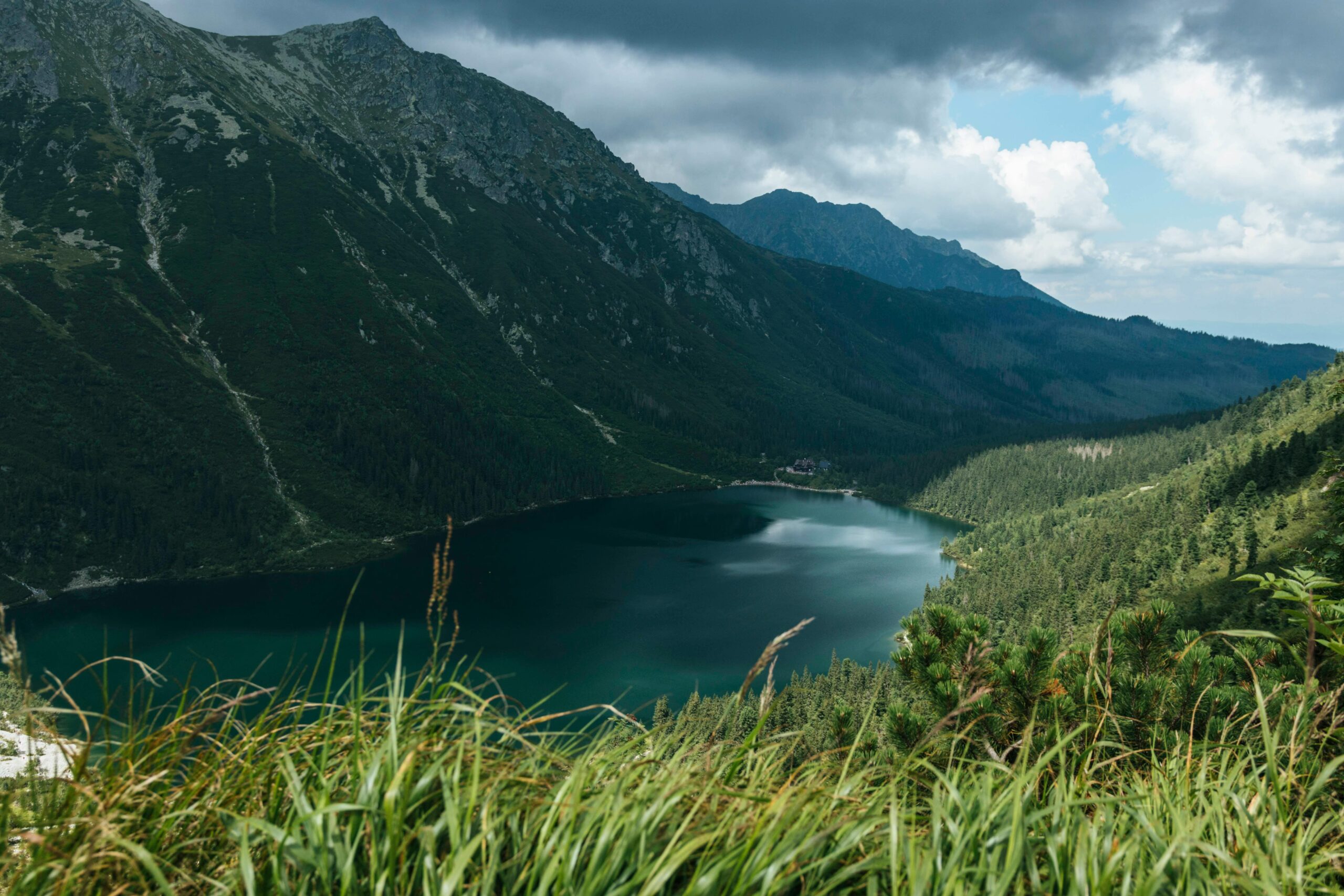 Spektakularny widok na górskie jezioro w Tatrach otoczone stromymi szczytami i lasami iglastymi. Dzika przyroda polskich gór z dramatycznym niebem - idealne miejsce na trekking i fotografię krajobrazową.