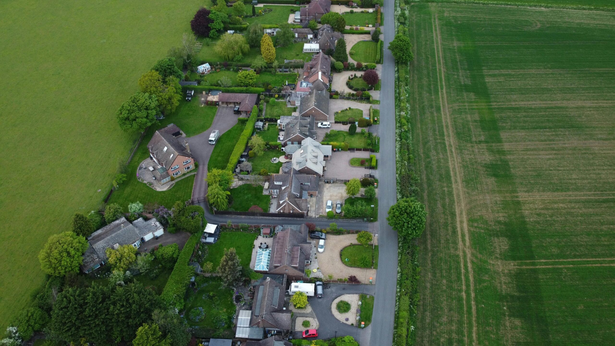 Panorama z lotu ptaka przedstawiająca spokojne osiedle domów jednorodzinnych z dużymi ogrodami, otoczone zielonymi polami uprawnymi i łąkami. Malowniczy krajobraz wiejski z nowoczesną zabudową mieszkaniową.