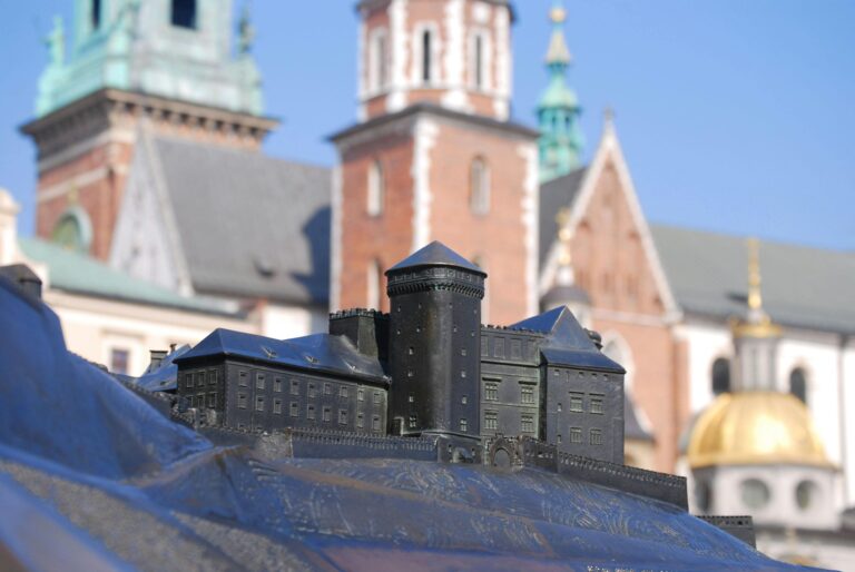 Miniaturowy model zamku na pierwszym planie z widokiem na krakowską katedrę z czerwonymi ceglanymi wieżami i zielonymi kopułami. Unikalne ujęcie łączące makietę z historyczną architekturą Krakowa w tle.