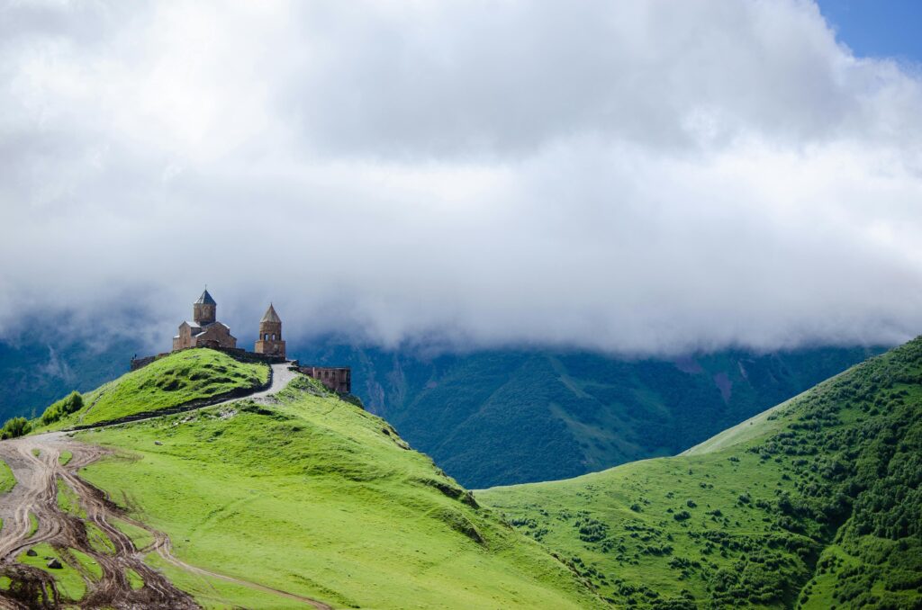 Panoramiczny widok na klasztor Gergeti położony na stromym zielonym wzgórzu w regionie Kazbegi w Gruzji - starożytna kamienną cerkiew z wieżami na tle majestycznych gór Kaukazu pokrytych mgłą i chmurami
