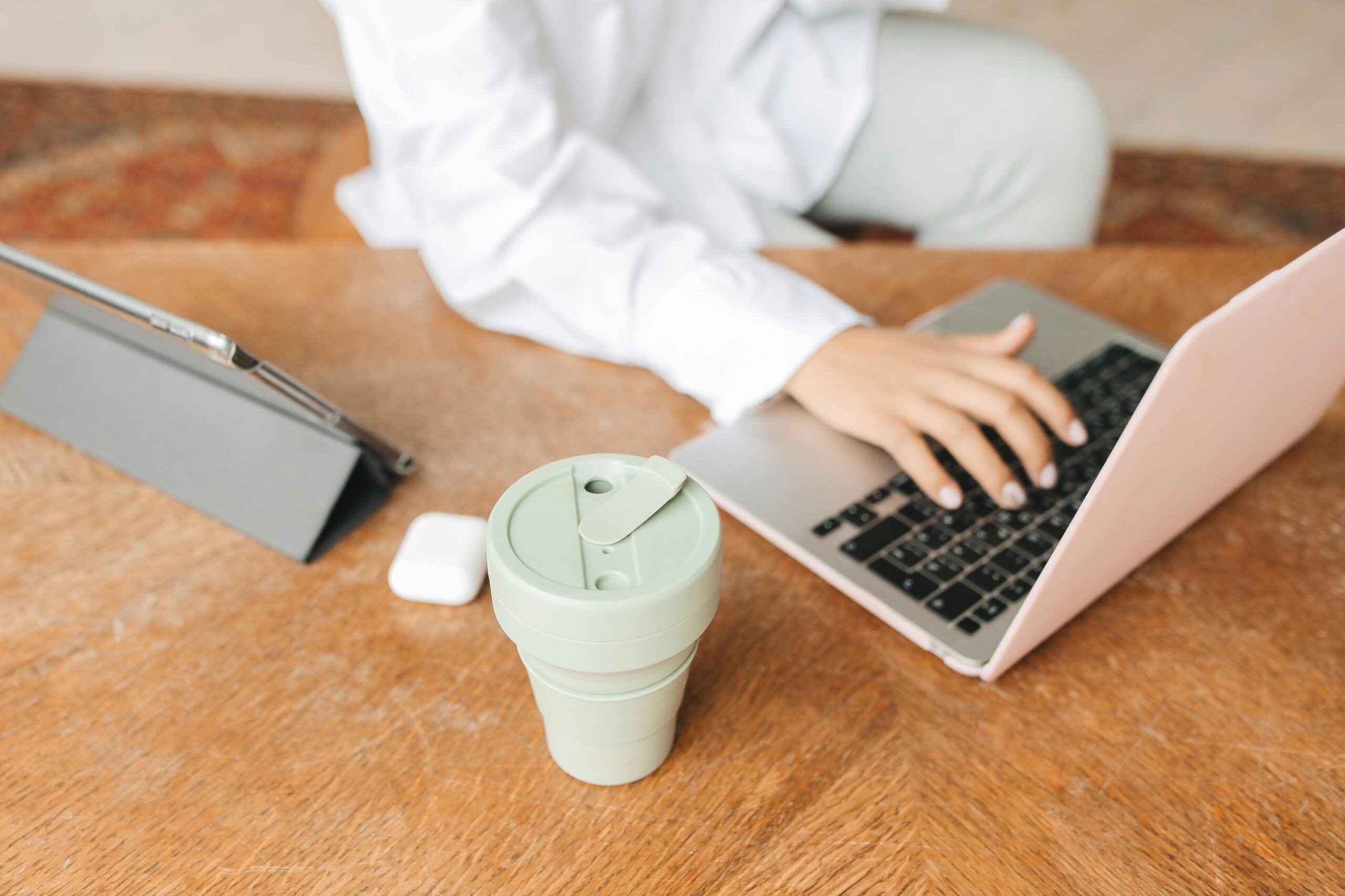 Praca zdalna przy laptopie - kobieta pracująca w home office przy drewnianym biurku z ekologicznym kubkiem termicznym wielokrotnego użytku i tabletem.