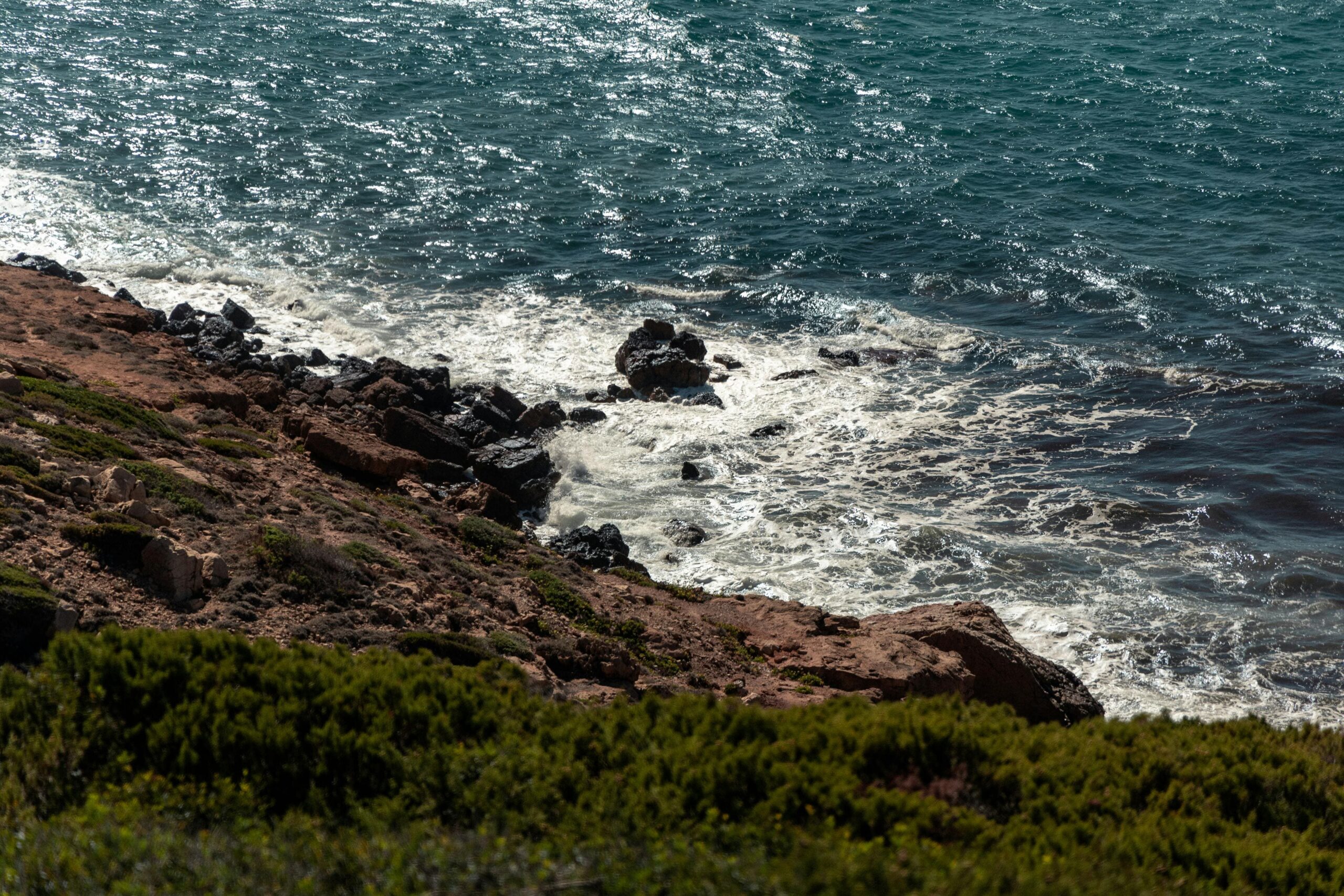 Skaliste wybrzeże Sardynia fale rozbijające się o skały - dzikie brzegi Morza Śródziemnego z ciemnym bazaltem, śródziemnomorska makchia na klifach, nieskażona natura włoskiego wybrzeża