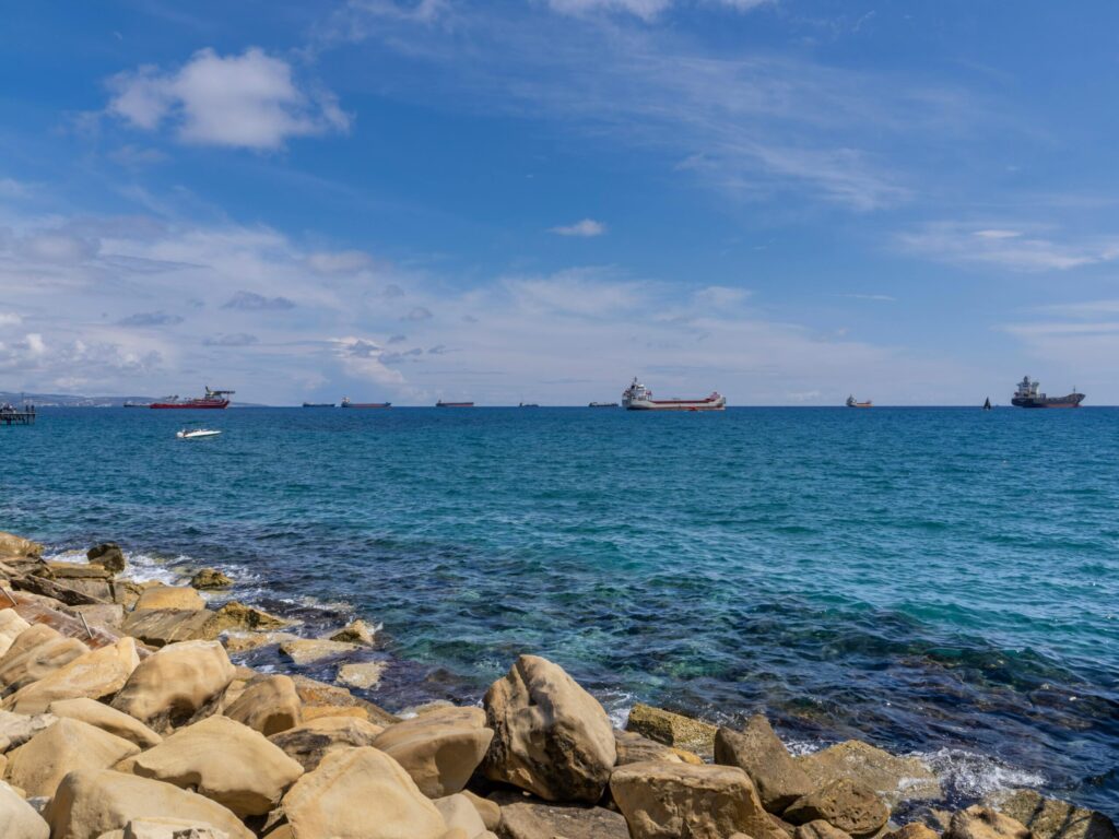 Cypr, Limassol - Morze Śródziemne wybrzeże ze skalistymi formacjami - turkusowe wody z widokiem na statki handlowe, naturalne wybrzeże z kamienistą plażą, panorama morska z błękitnym niebem i chmurami