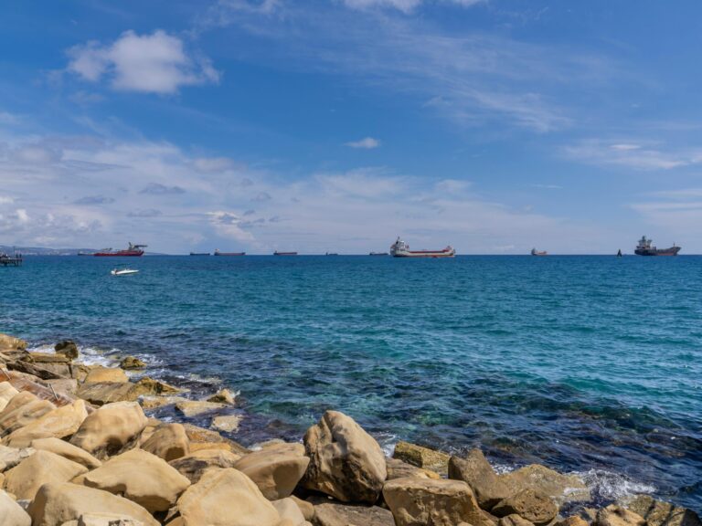 Cypr, Limassol - Morze Śródziemne wybrzeże ze skalistymi formacjami - turkusowe wody z widokiem na statki handlowe, naturalne wybrzeże z kamienistą plażą, panorama morska z błękitnym niebem i chmurami